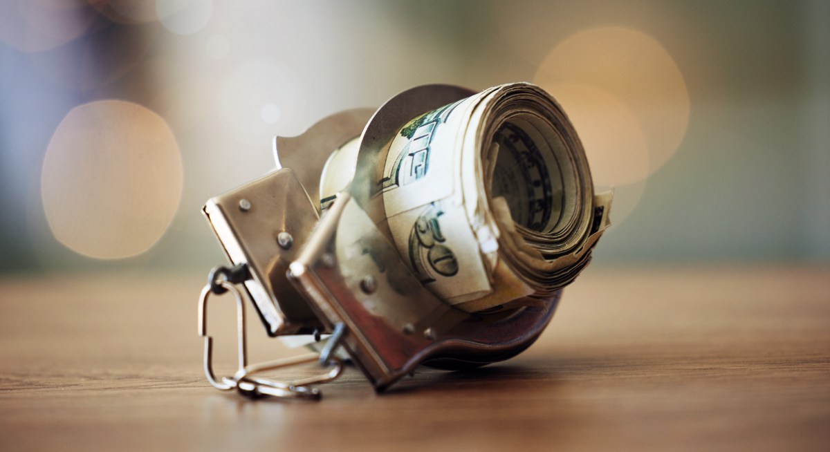 A photograph of a roll of dollar bills inside a pair of handcuffs, on a wooden tabletop